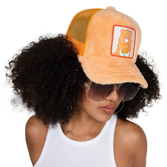 Woman wearing orange faux fur trucker cap featuring a woven patch of Friendship Bear from Care Bears attached to the crown.