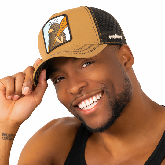 Man wearing light brown twill trucker cap with black mesh featuring a woven patch of Captain Caveman from the classic Hanna-Barbera cartoons attached to the crown.