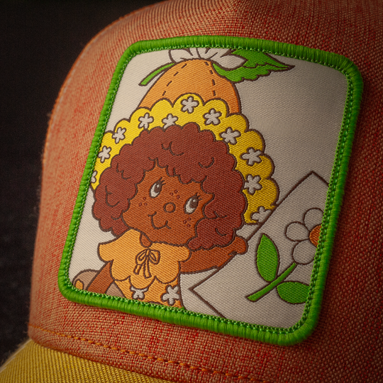 Close-up of a woven patch featuring an illustration of Orange Blossom character from Strawberry Shortcake on orange linen trucker cap hat