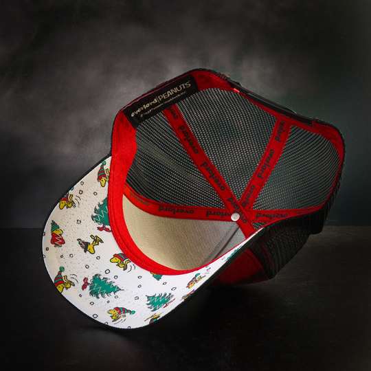 Inside view of red and black buffalo plaid flannel trucker cap with Woodstock in a beanie and Christmas trees scattered on white underbrim.