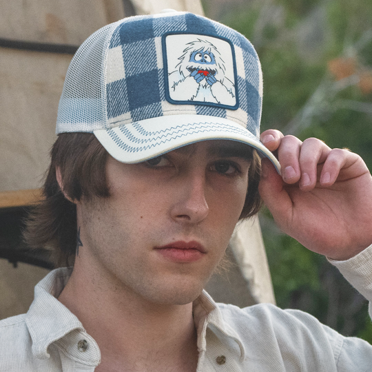 Man wearing a navy and white plaid trucker cap with white mesh featuring a woven patch of Bumble from Rudolph the Red Nosed Reindeer on the crown.