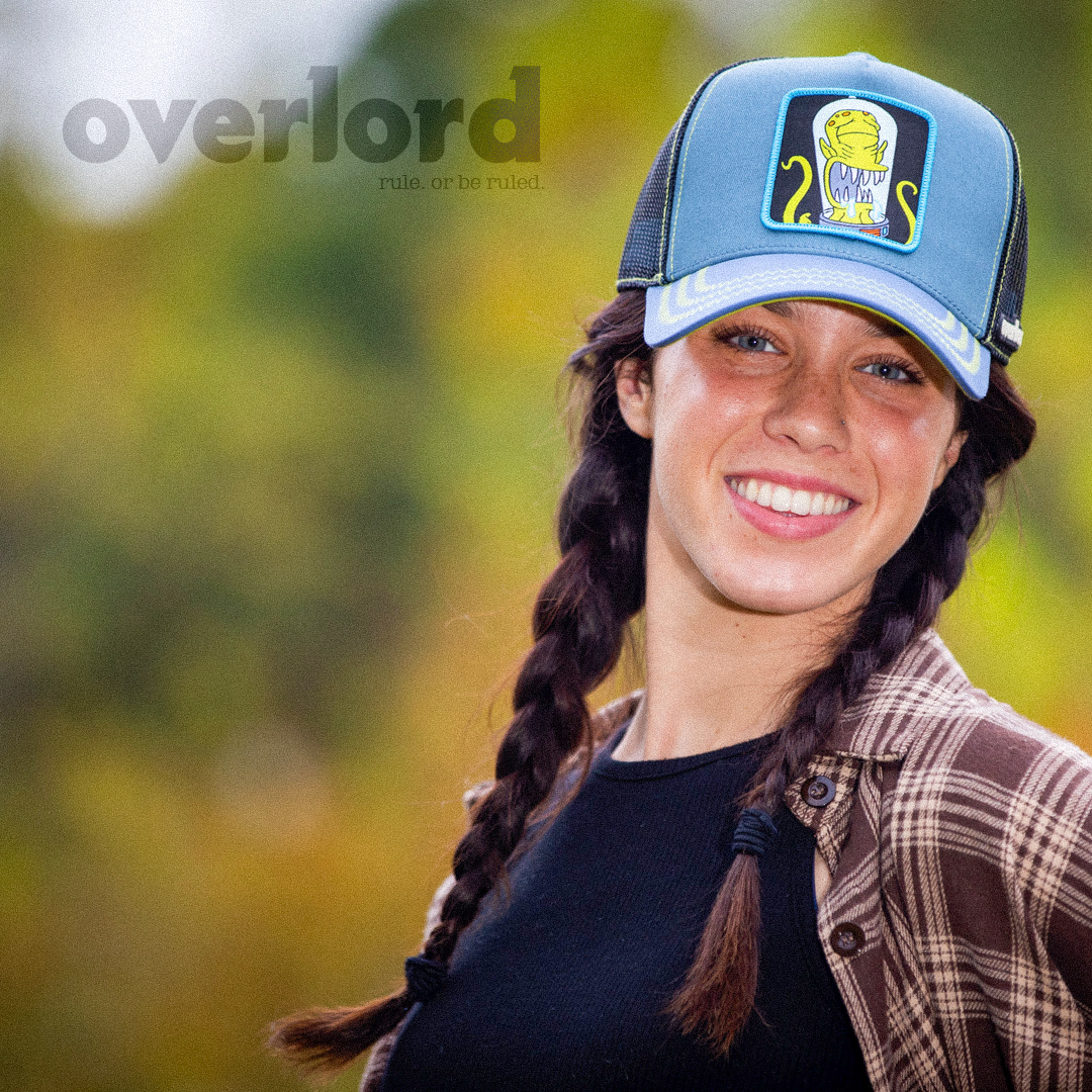 Woman wearing blue OVERLORD X The Simpsons present Kodos the alien trucker baseball cap hat with lime green zig zag stitching. PVC Overlord logo.