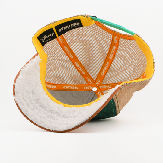 Underside of the Bashful from Disney's Snow White and the Seven Dwarfs retro trucker cap showing a white fuzzy-textured brim, a Disney tag, tan mesh crown, and orange Overlord branded taping.