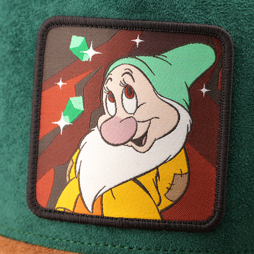 Woven patch of Bashful from Disney's Snow White and the Seven Dwarfs smiling shyly against a mine and gem-themed background, with a black border, stitched onto the green suede-textured retro trucker cap.