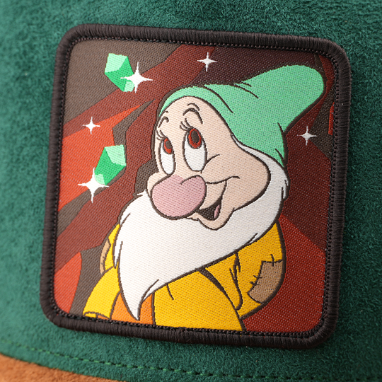 Woven patch of Bashful from Disney's Snow White and the Seven Dwarfs smiling shyly against a mine and gem-themed background, with a black border, stitched onto the green suede-textured retro trucker cap.