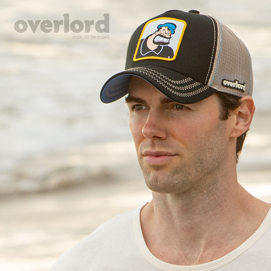 Man wearing black OVERLORD X Popeye angry Brutus Bluto trucker baseball cap hat with khaki zig zag stitching. PVC Overlord logo.