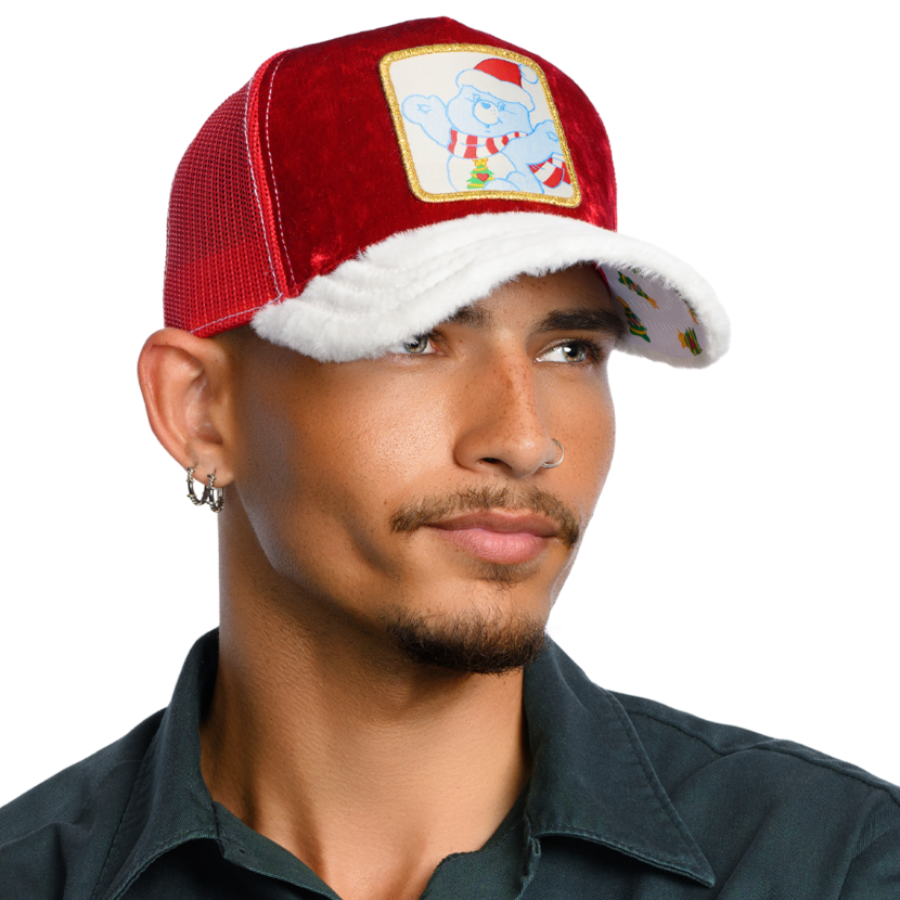 Man wearing red crushed velvet trucker cap with white faux fur brim and red mesh featuring a woven patch of Christmas Wishes Bear from Care Bears wearing a Santa hat and scarf attached to the crown.