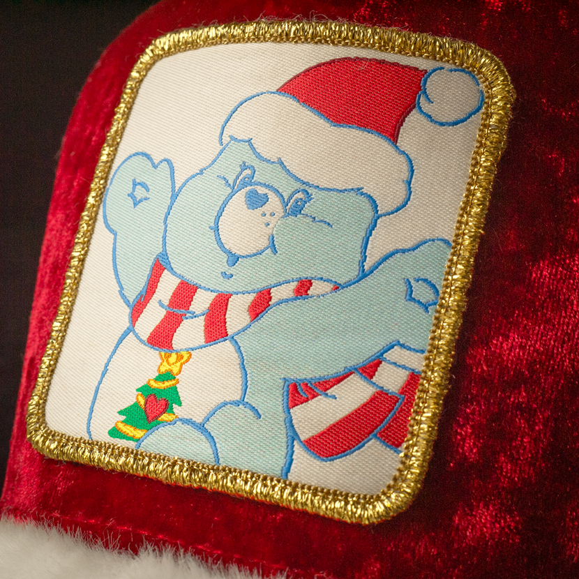 Close-up of a woven patch of Christmas Wishes Bear from Care Bears wearing a Santa hat and scarf attached to the crown.