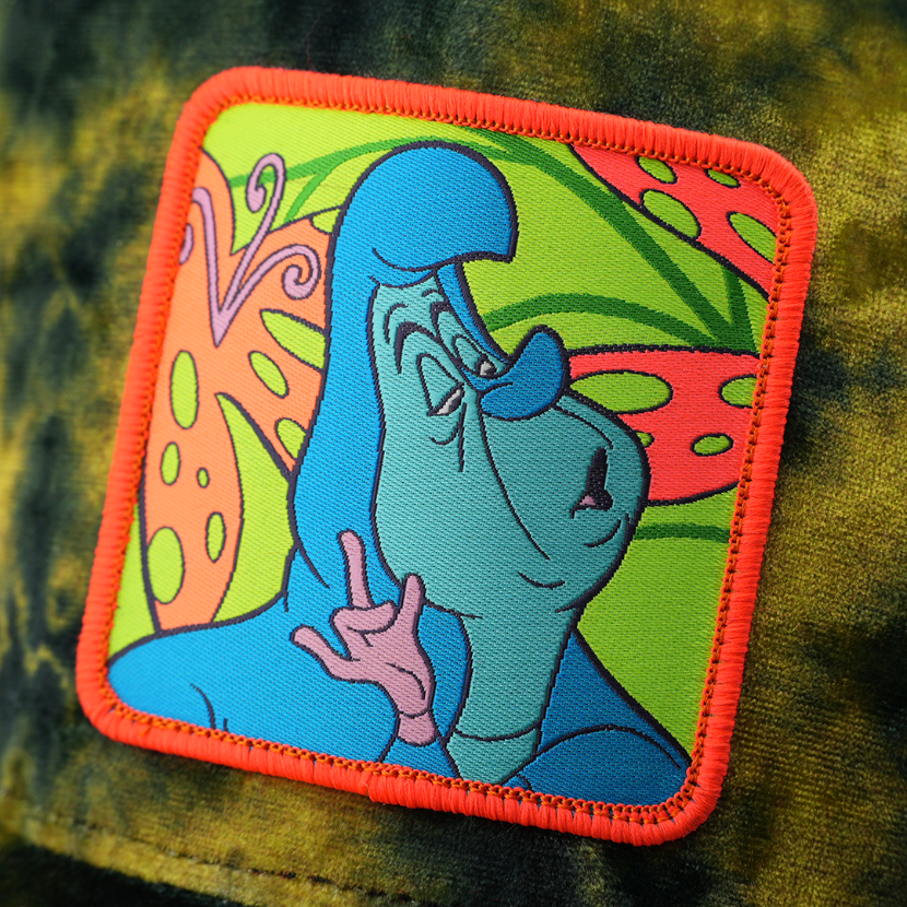 Woven patch of the Caterpillar from Disney's Alice in Wonderland pointing on a colorful mushroom-print background, with red border, stitched onto the green and black tie-dye-textured retro trucker cap.