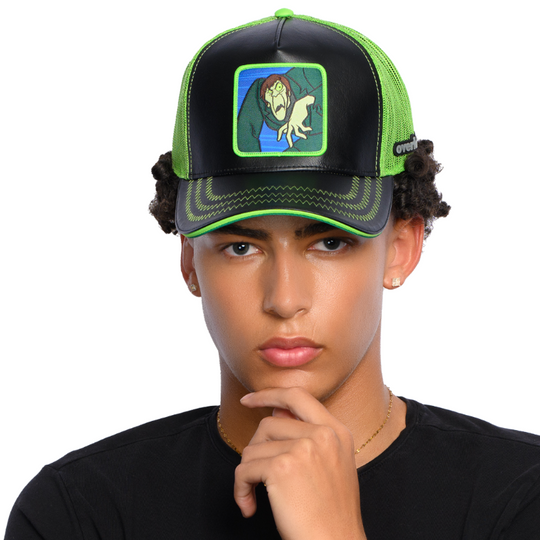 Man wearing black faux leather trucker cap with green mesh featuring a woven patch of the Creeper villain from Scooby Doo attached to the crown.