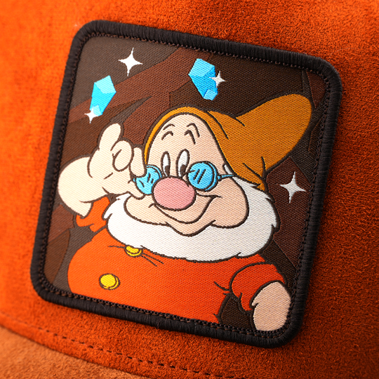Woven patch of Doc from Disney's Snow White and the Seven Dwarfs adjusting his glasses against a mine and gem-themed background, with a black border, stitched onto the orange suede-textured retro trucker cap.