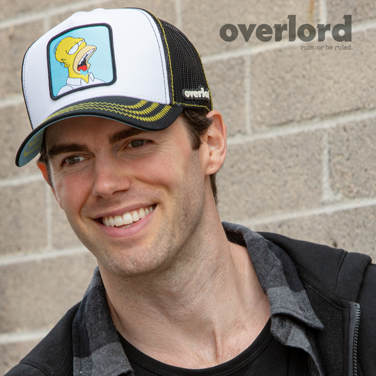 Man wearing white and black OVERLORD X The Simpsons Homer drooling trucker baseball cap hat with yellow zig zag stitching. PVC Overlord logo.