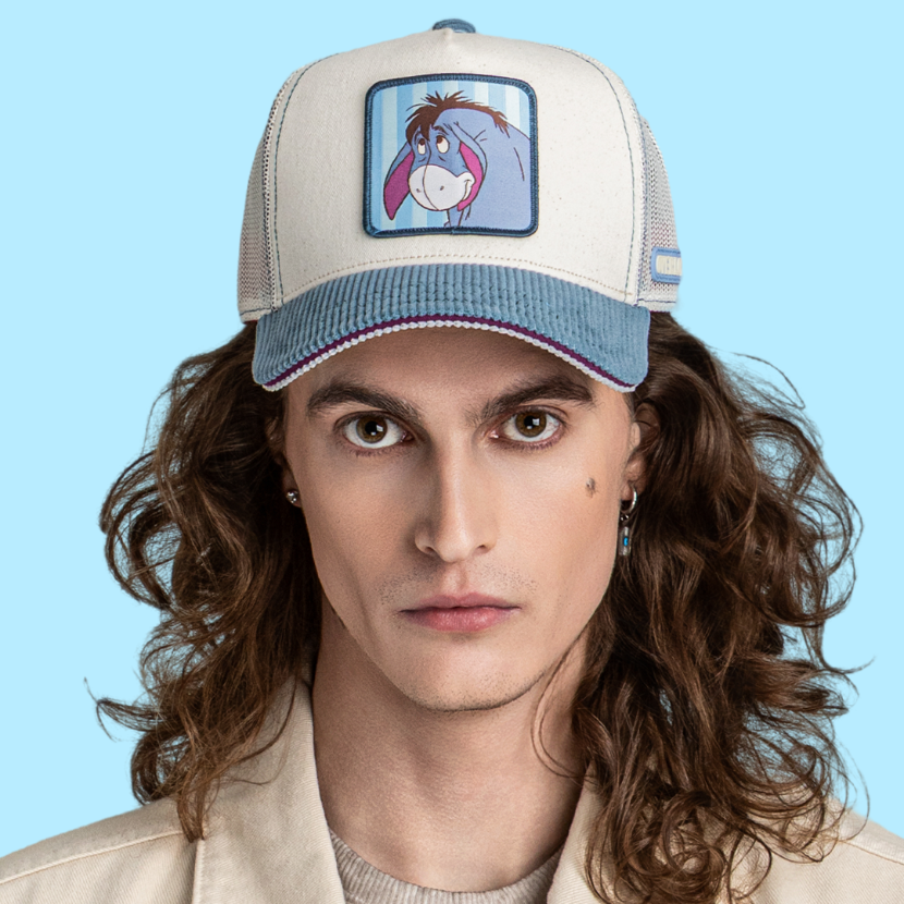 Model wearing the off-white Eeyore retro trucker cap with blue brim and off-white mesh sides.