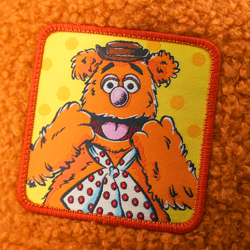 Close-up of a woven patch showing Fozzie Bear from The Muppets in a bowler hat and polka-dot bow tie on a yellow polka-dot background, stitched onto boucle-textured burnt orange fabric with a red border.