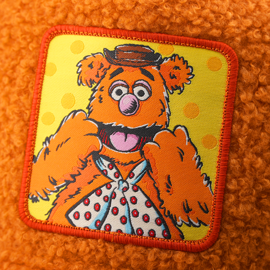 Close-up of a woven patch showing Fozzie Bear from The Muppets in a bowler hat and polka-dot bow tie on a yellow polka-dot background, stitched onto boucle-textured burnt orange fabric with a red border.