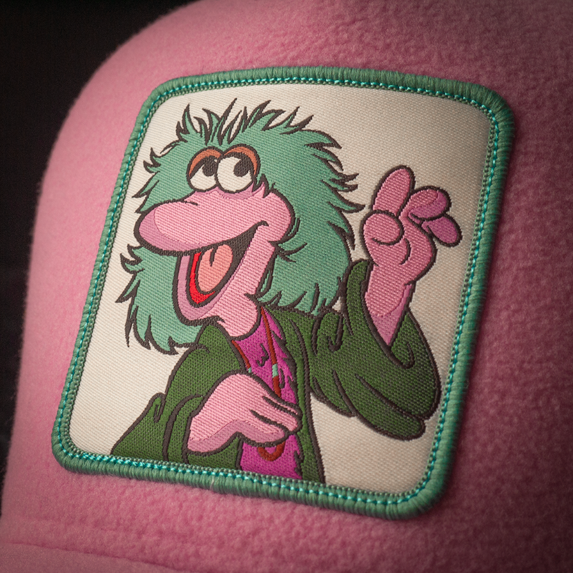 A trucker cap with a woven patch featuring the character Mokey from Fraggle Rock with a serene smile and flower-child hair in soft lavender tones with rainbow-stitched mesh.