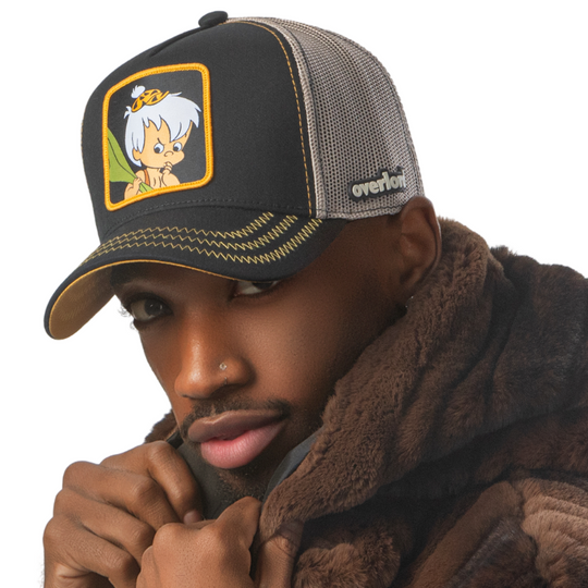 Man wearing black OVERLORD X Flintstones Bamm Bamm trucker baseball cap with yellow zig zag stitching. PVC Overlord logo.