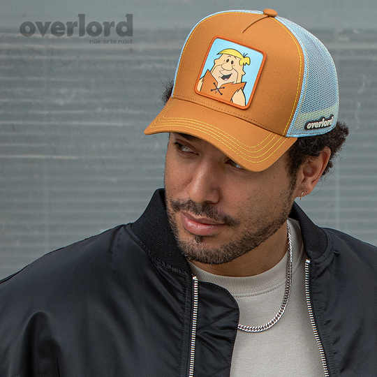 Man wearing brown OVERLORD X Flintstones Barney Rubble trucker baseball cap with yellow stitching. PVC Overlord logo.