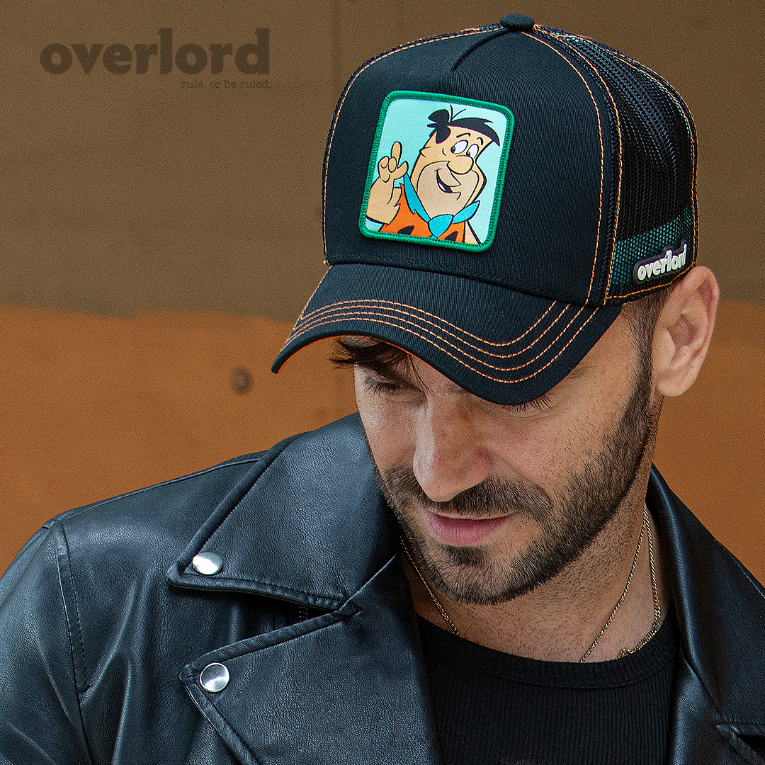Man wearing black OVERLORD X Flintstones Fred Flintstone trucker baseball cap with orange stitching. PVC Overlord logo.