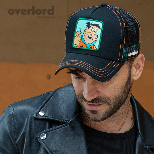 Man wearing black OVERLORD X Flintstones Fred Flintstone trucker baseball cap with orange stitching. PVC Overlord logo.