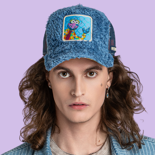 Model wearing the fuzzy-textured blue Gonzo from The Muppets retro trucker cap with a mustard yellow-edged brim and dark mesh side.