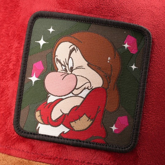 Woven patch of Grumpy from Disney's Snow White and the Seven Dwarfs frowning with arms crossed against a mine and gem-themed background, with a black border, stitched onto the red suede-textured retro trucker cap.