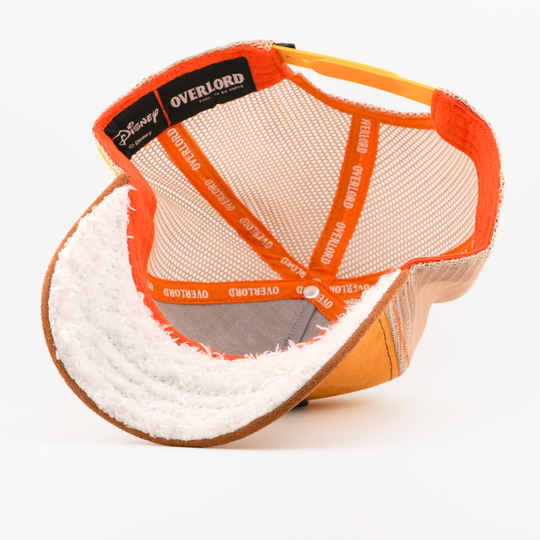 Underside of the Happy from Disney's Snow White and the Seven Dwarfs retro trucker cap showing a white fuzzy-textured brim, a Disney tag, tan mesh crown, and orange Overlord branded taping.