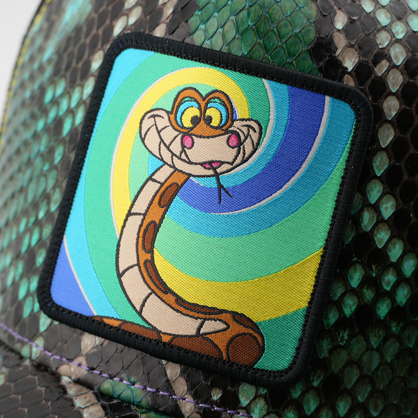 Close-up woven patch of Kaa from Disney's The Jungle Book with a hypnotic grin, on a swirl-pattern background with a black border, stitched onto a green snakeskin-textured retro trucker cap.