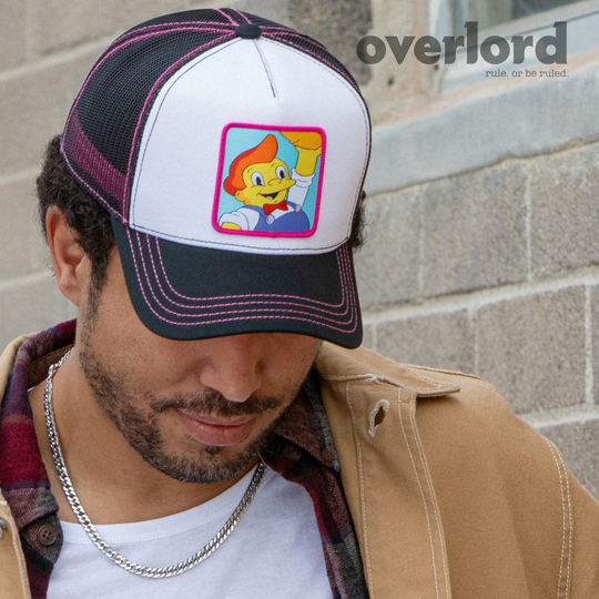 Man wearing white and black OVERLORD X The Simpsons Lard Lad holding donut trucker baseball cap hat with pink stitching. PVC Overlord logo.