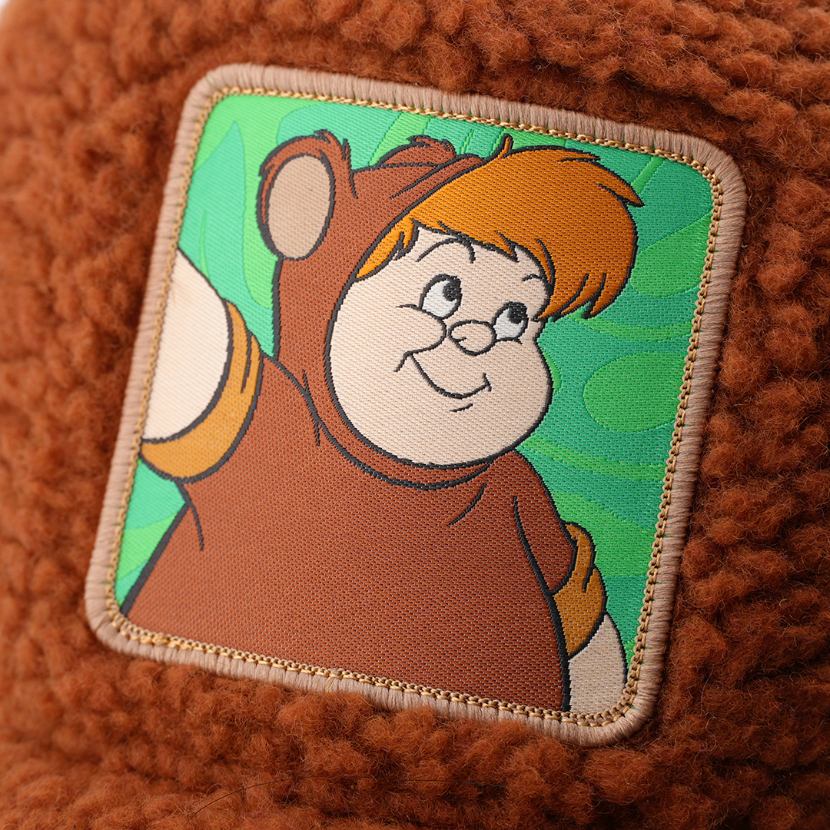 Close-up of a woven patch showing Lost Boy Cubby from Disney's Peter Pan in a bear costume on a green background, stitched onto fuzzy-textured brown fabric with a tan border.