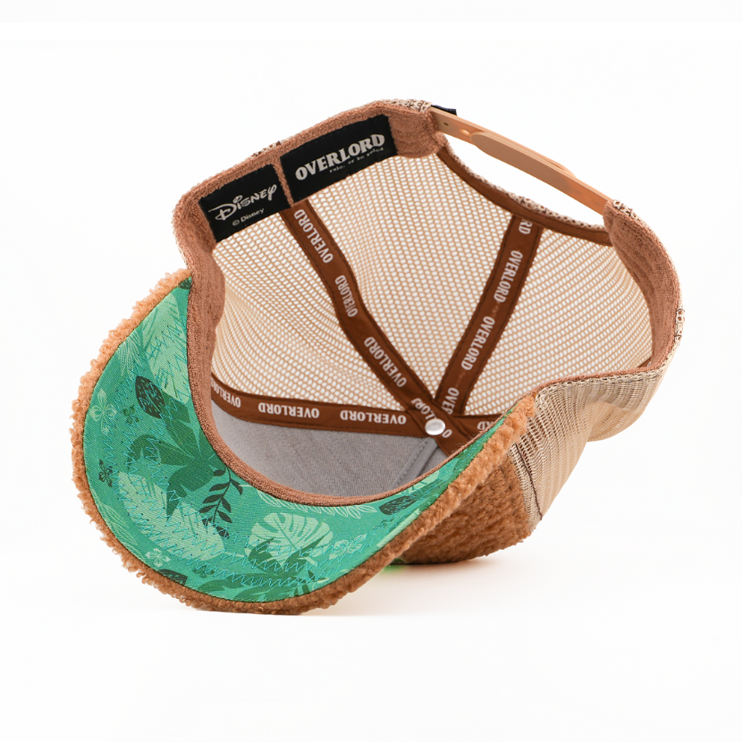 Underside of the Lost Boy Nibs retro trucker cap, showing a green tropical leaf-print brim, a Disney tag, tan mesh crown, and brown Overlord branded taping with white text.