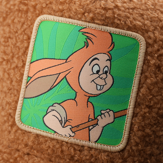 Close-up of a woven patch showing Lost Boy Nibs from Disney's Peter Pan holding a stick on a green leaf background, stitched onto fuzzy-textured brown fabric with a tan border.