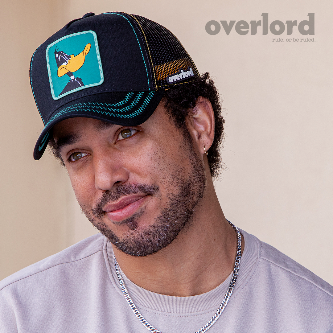 Man wearing black OVERLORD X Looney Tunes Daffy Duck trucker baseball cap hat with teal zig zag stitching. PVC Overlord logo.