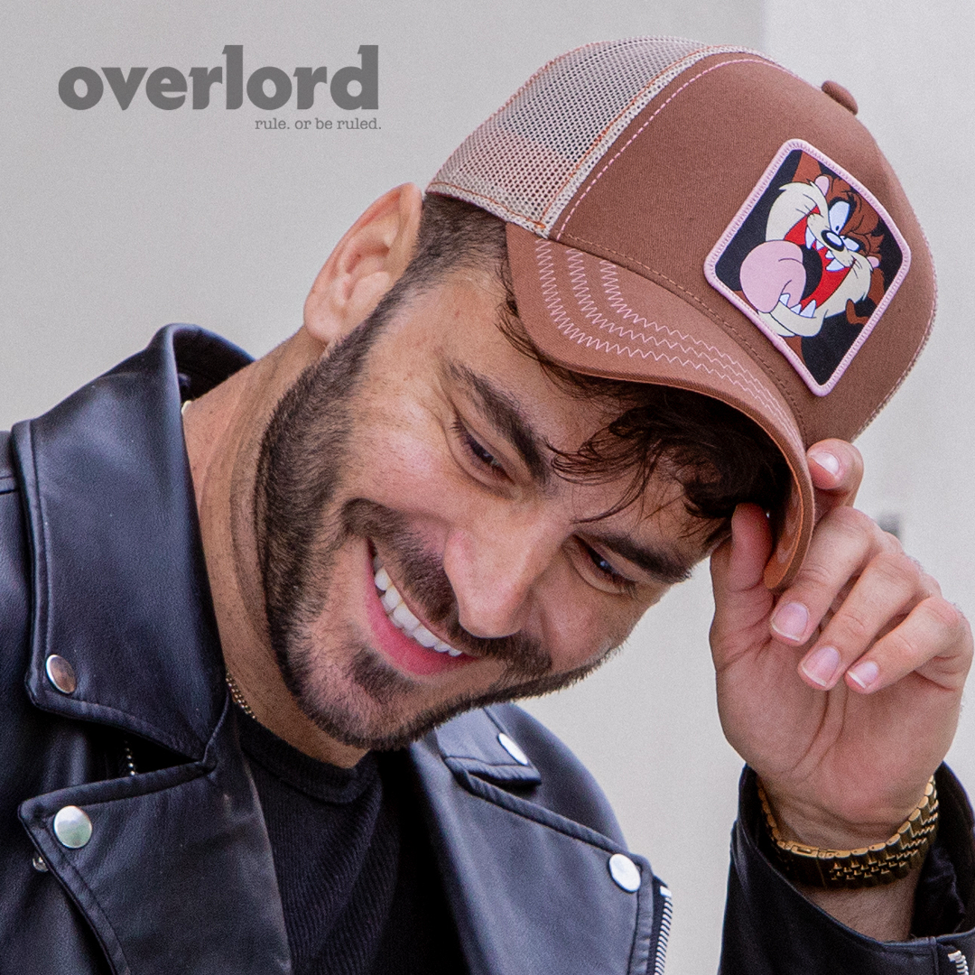 Man wearing brown OVERLORD X Looney Tunes smiling Tasmanian Devil trucker baseball cap hat with pink zig zag stitching. PVC Overlord logo.