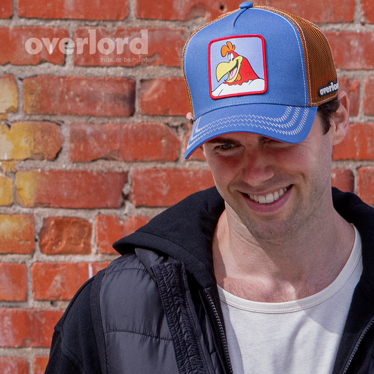 Man wearing blue OVERLORD X Looney Tunes smiling Foghorn trucker baseball cap with cream stitching. PVC Overlord logo.