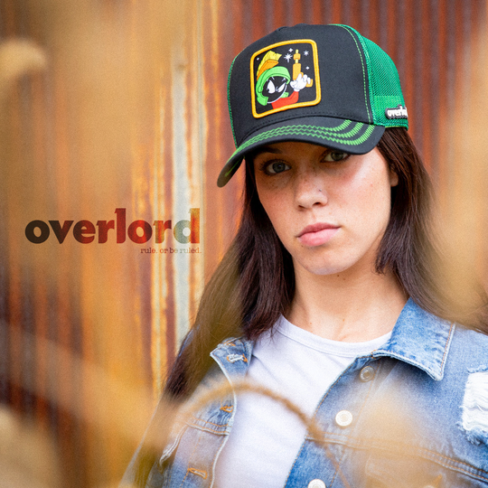 Woman wearing black OVERLORD X Looney Tunes Marvin the Martian holding a laser gun trucker baseball cap hat with green zig zag stitching. PVC Overlord logo.
