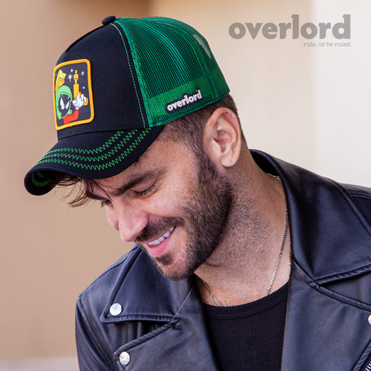 Man wearing black OVERLORD X Looney Tunes Marvin the Martian holding a laser gun trucker baseball cap hat with green zig zag stitching. PVC Overlord logo.