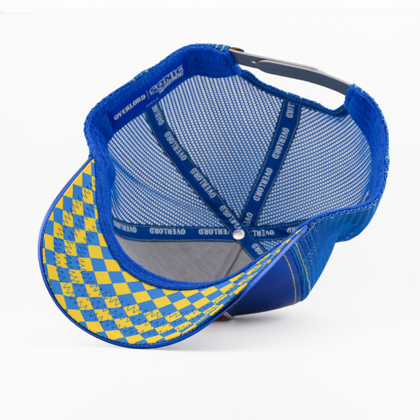 Underside of the Metal Sonic from Sonic the Hedgehog retro trucker cap, showing a blue and yellow argyle-pattern brim, Sonic the Hedgehog tag, grey mesh crown, and blue Overlord branded taping with yellow text.