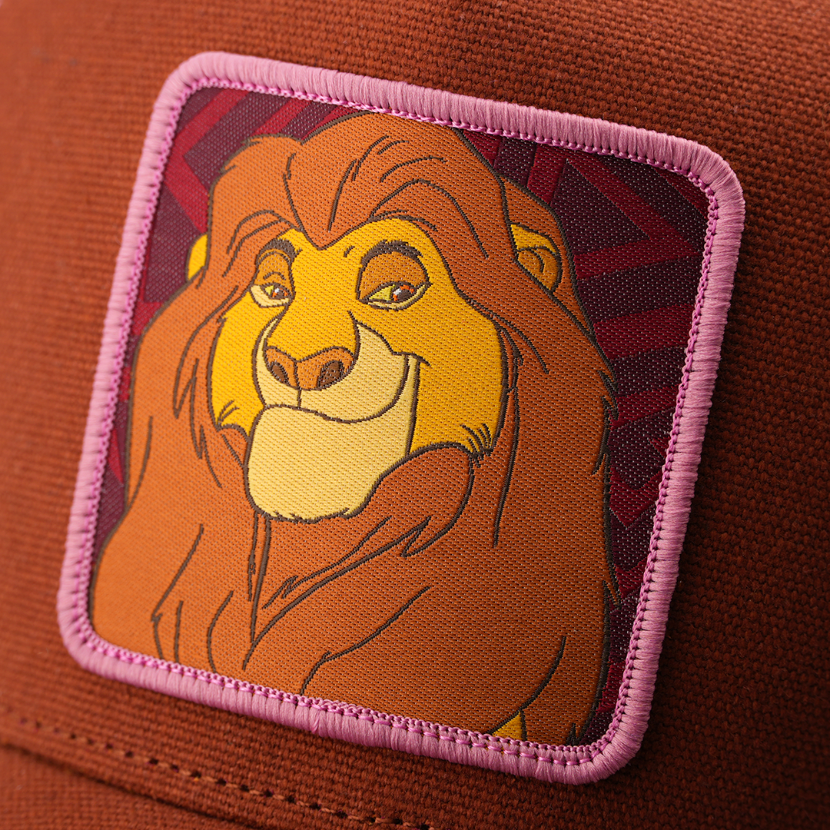 Close-up woven patch of Mufasa from Disney's The Lion King smiling warmly, on a dark red patterned background with a pink border, stitched onto a brown corduroy-textured retro trucker cap.