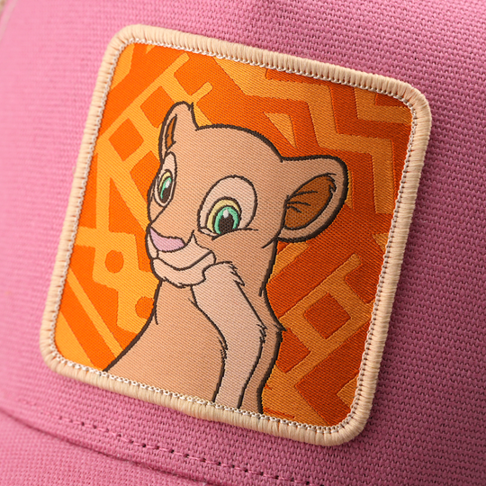 Close-up woven patch of young Nala from Disney's The Lion King with a soft smile, on an orange patterned background with a tan border, stitched onto a pink corduroy-textured retro trucker cap.