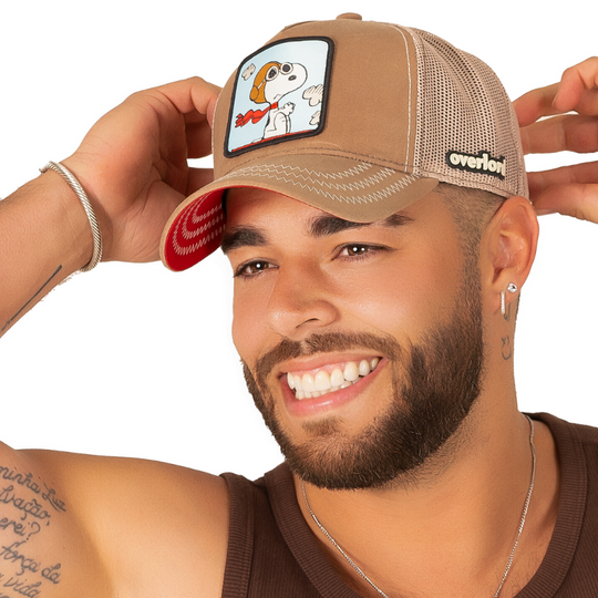 Man wearing brown waxed canvas trucker cap with khaki mesh featuring a woven patch of Snoopy Flying Ace from the Peanuts Comics attached to the crown.