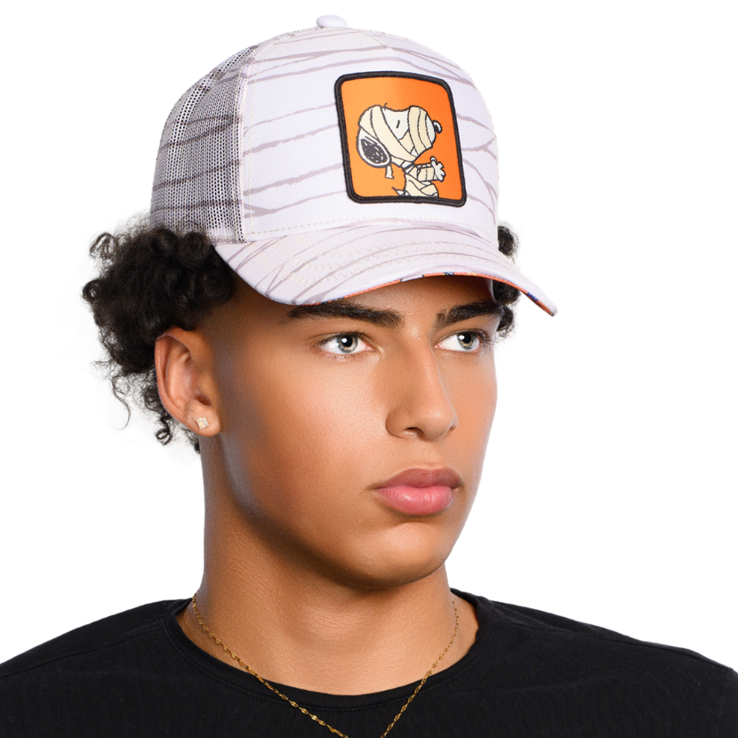 Man wearing cream colored trucker cap with printed mummy wraps on the crown, brim and mesh featuring a woven patch of Mummy Snoopy on the crown.