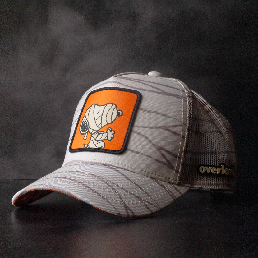Cream colored trucker cap with printed mummy wraps on the crown, brim and mesh featuring a woven patch of Mummy Snoopy on the crown.