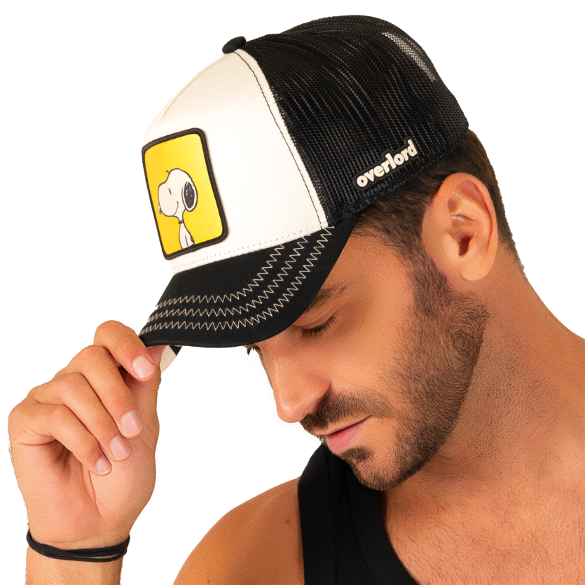 Man wearing black and white twill trucker cap with black mesh featuring a woven patch of Snoopy from the Peanuts Comics on the crown.