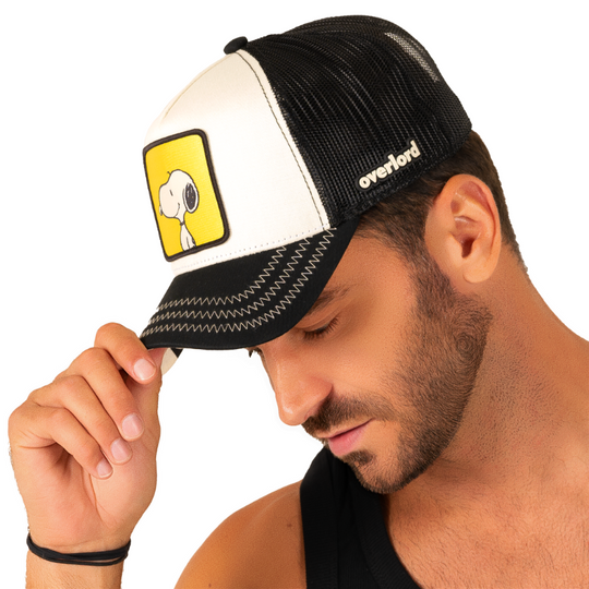 Man wearing black and white twill trucker cap with black mesh featuring a woven patch of Snoopy from the Peanuts Comics on the crown.