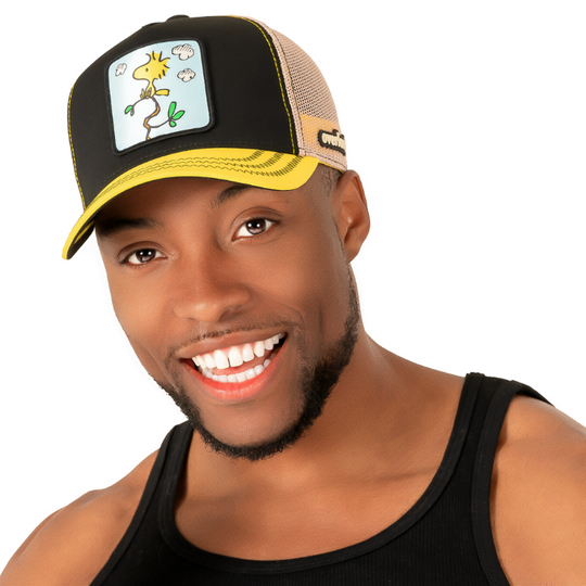 Man wearing black and yellow twill trucker cap with khaki mesh featuring a woven patch of Woodstock from Peanuts on the crown.