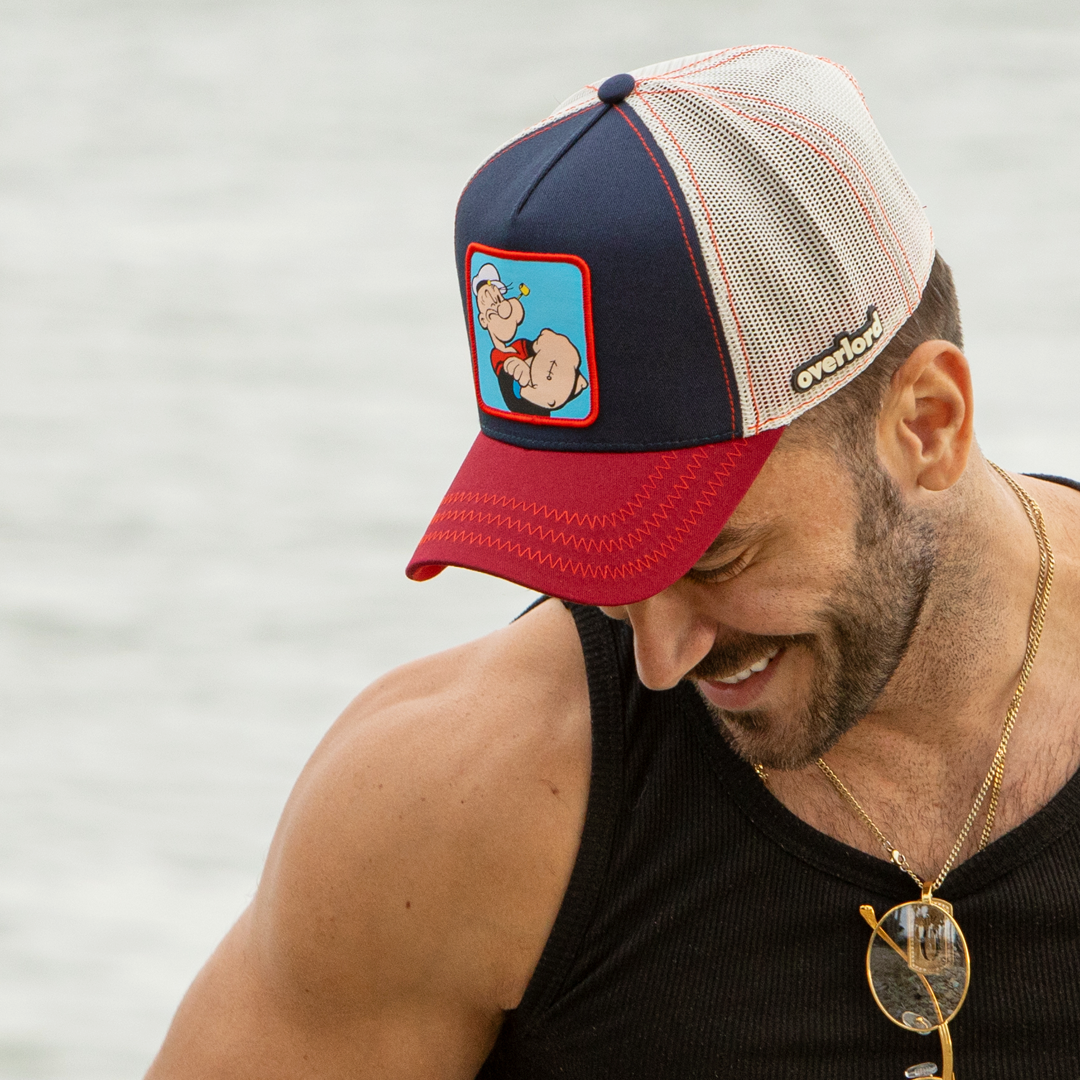 Man wearing navy and red OVERLORD X Popeye smug Popeye trucker baseball cap hat with red zig zag stitching. PVC Overlord logo.
