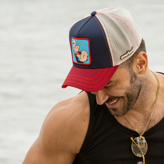 Man wearing navy and red OVERLORD X Popeye smug Popeye trucker baseball cap hat with red zig zag stitching. PVC Overlord logo.
