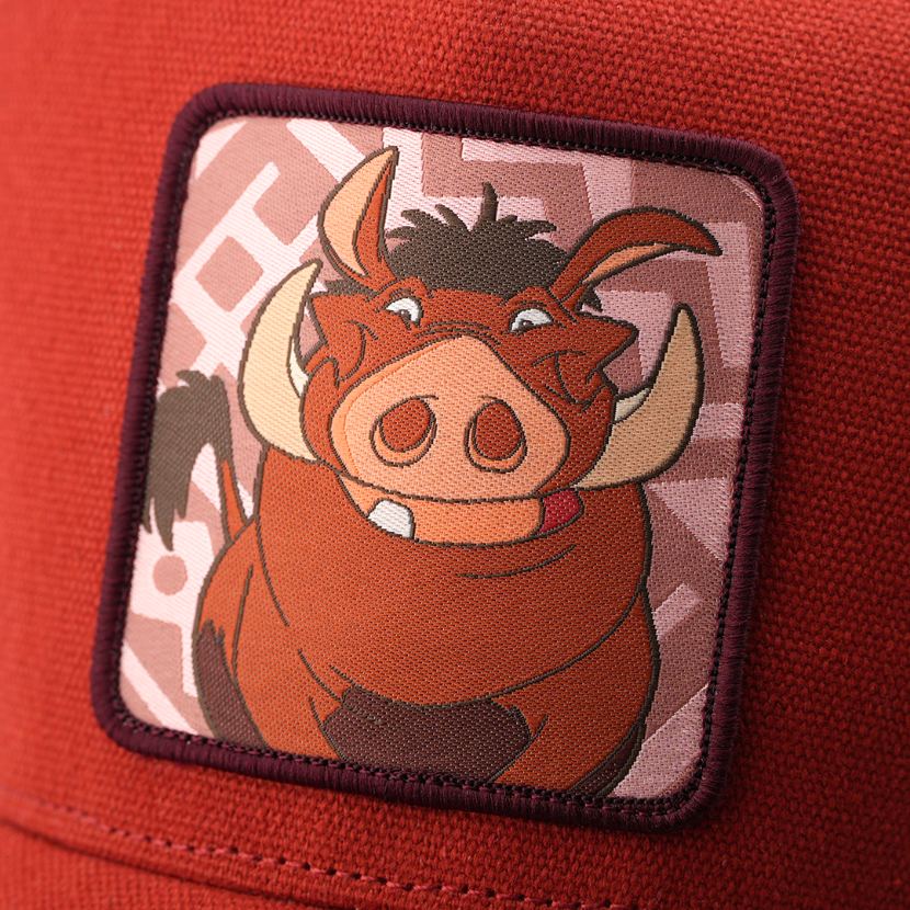Close-up woven patch of Pumbaa from Disney's The Lion King smiling with tongue out, on a pink patterned background with a dark purple border, stitched onto a rust-red corduroy-textured retro trucker cap.