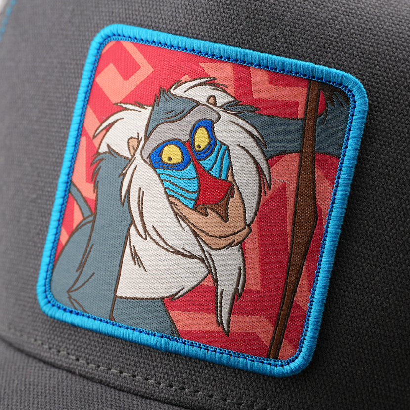 Close-up woven patch of Rafiki from Disney's The Lion King holding his staff, on a red patterned background with a blue border, stitched onto a grey corduroy-textured retro trucker cap.